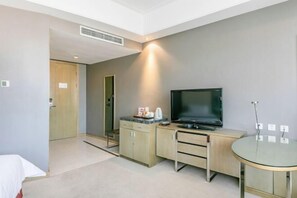 A modern hotel room with a bed, a desk with a flat-screen TV, a small table, and a kitchenette area.