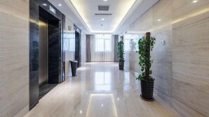 Un couloir d’hôtel moderne, avec un sol en marbre, un ascenseur central et des plantes en pot.