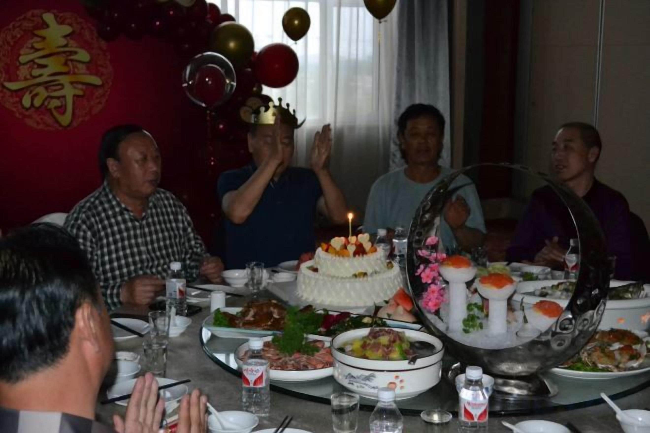 A group of people seated around a table with a cake and various dishes, in a room with a red wall and a decorative emblem.
