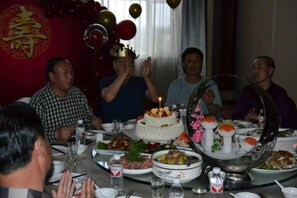 A group of people seated around a table with a cake and various dishes, in a room with a red wall and a decorative emblem. - Panshan Yinlong Hotel (Panshan)