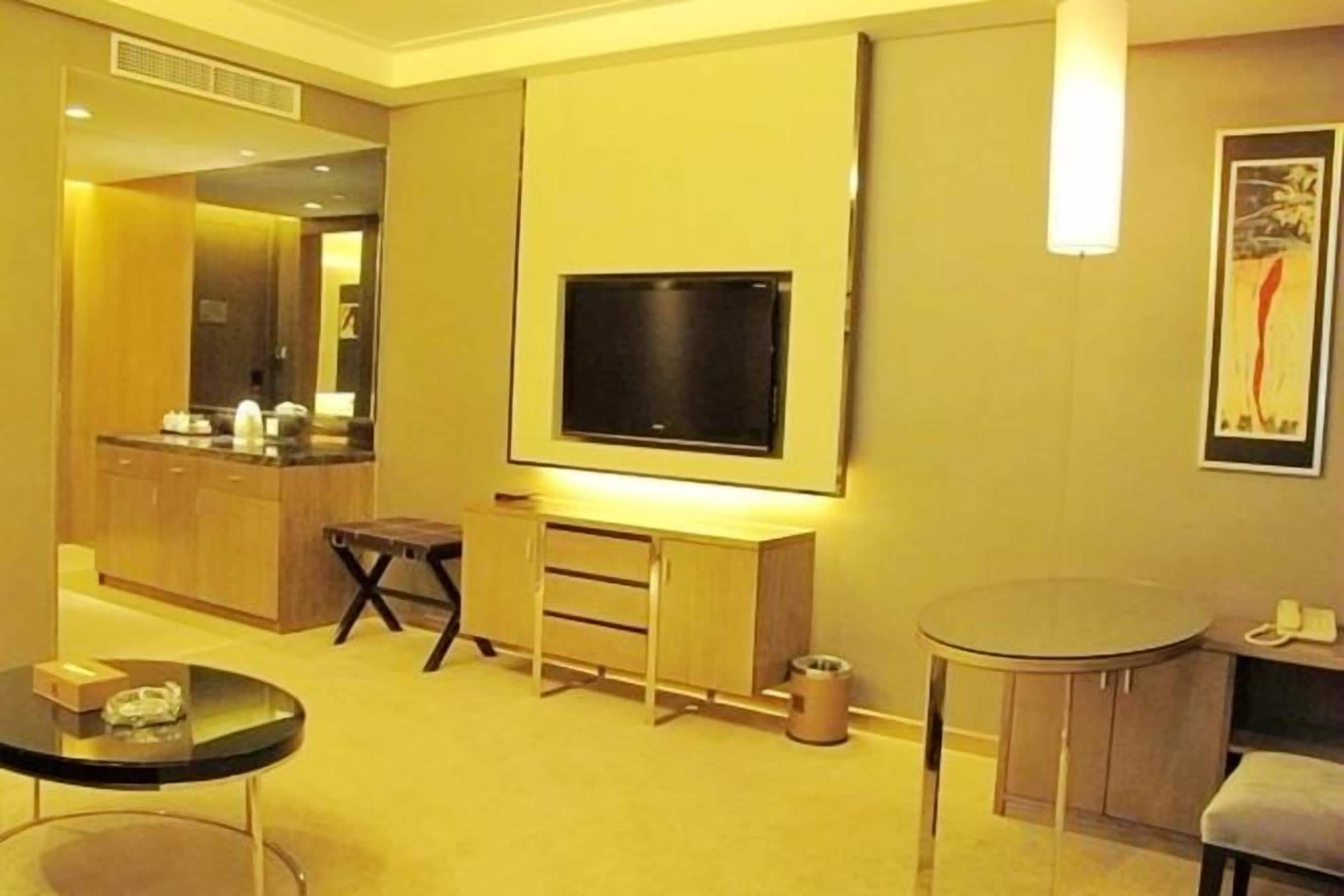 A hotel room with a flat-screen TV mounted on the wall, a small round table, a seating area with a chair, and a kitchenette in the background.