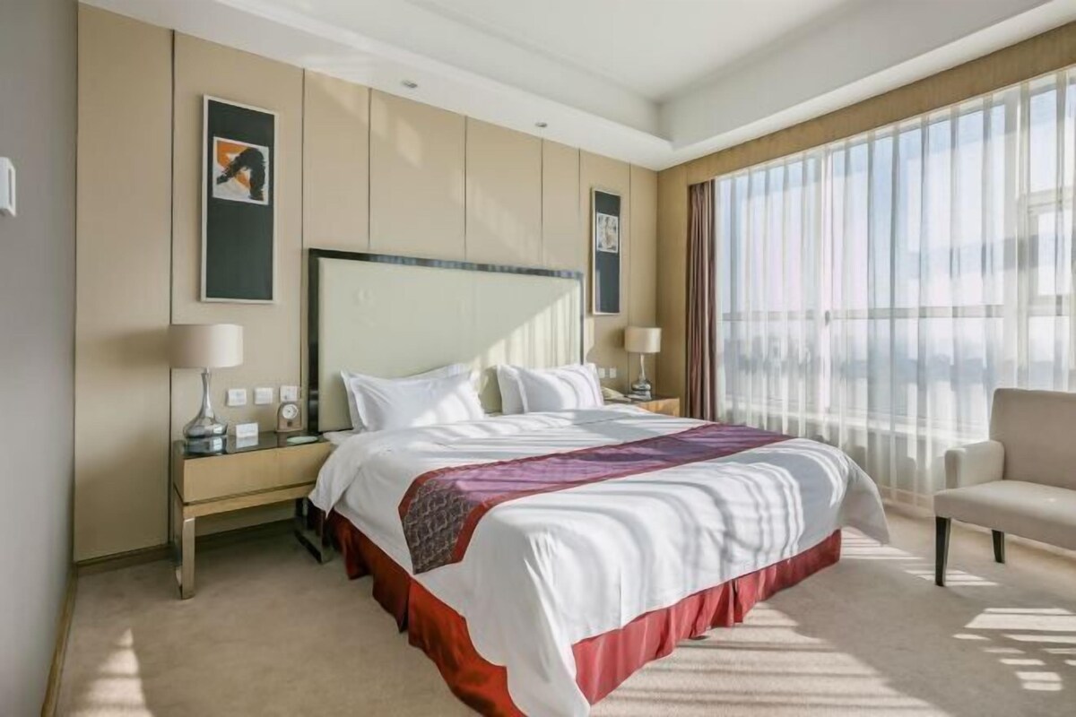 A hotel room with a large bed, bedside tables, a chair, and a large window with sheer curtains.