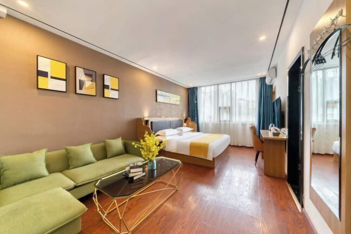Habitación de hotel moderna con un sofá verde, una cama con edredón blanco y amarillo, un escritorio de madera pequeño y un ventanal con cortinas.