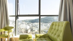 Quarto com uma grande janela com vista para a cidade, duas poltronas estofadas em verde e piso de madeira.