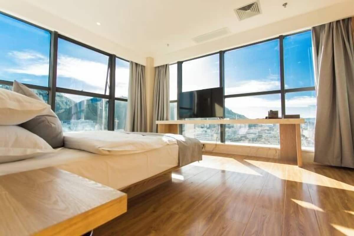 A modern hotel room with a large bed, a TV, and a view of a cityscape through large windows.