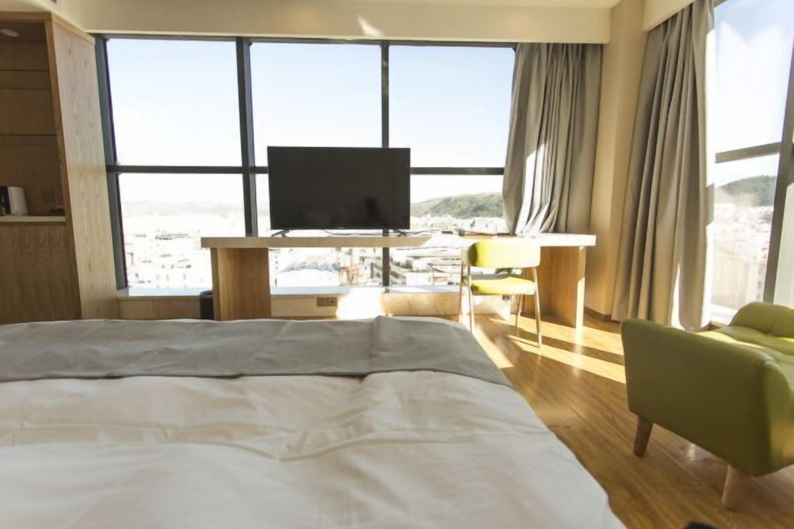 A hotel room with a bed, a desk with a TV, and a view of a cityscape through large windows.