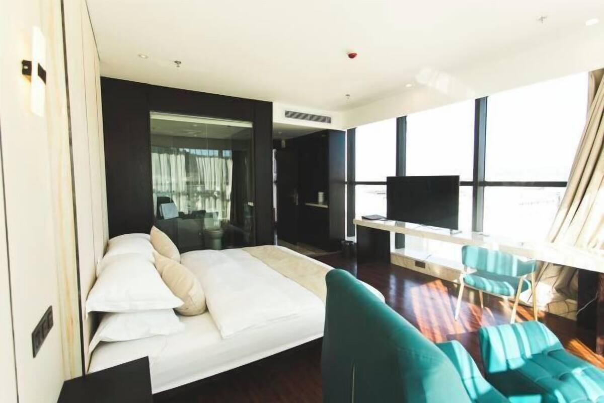 A modern hotel room with a bed, a TV, a desk, and a large window.