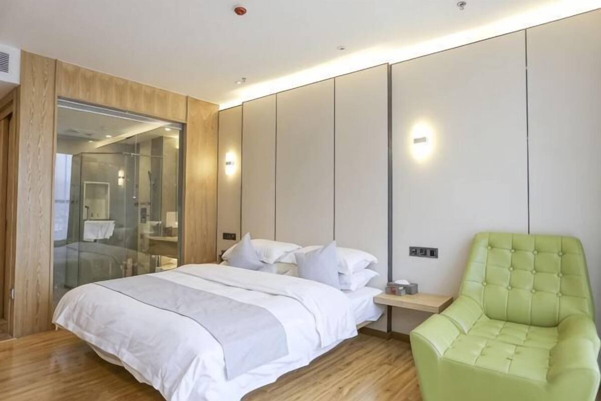 A modern hotel room with a bed, a green armchair, a glass shower enclosure, and a wooden floor.