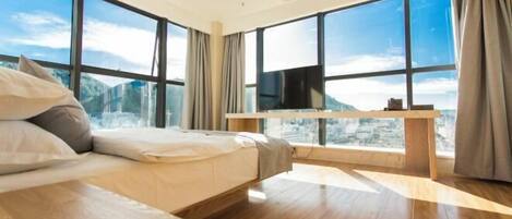 Una habitación de hotel moderna con una cama grande, un televisor y vistas a un paisaje urbano a través de amplias ventanas.