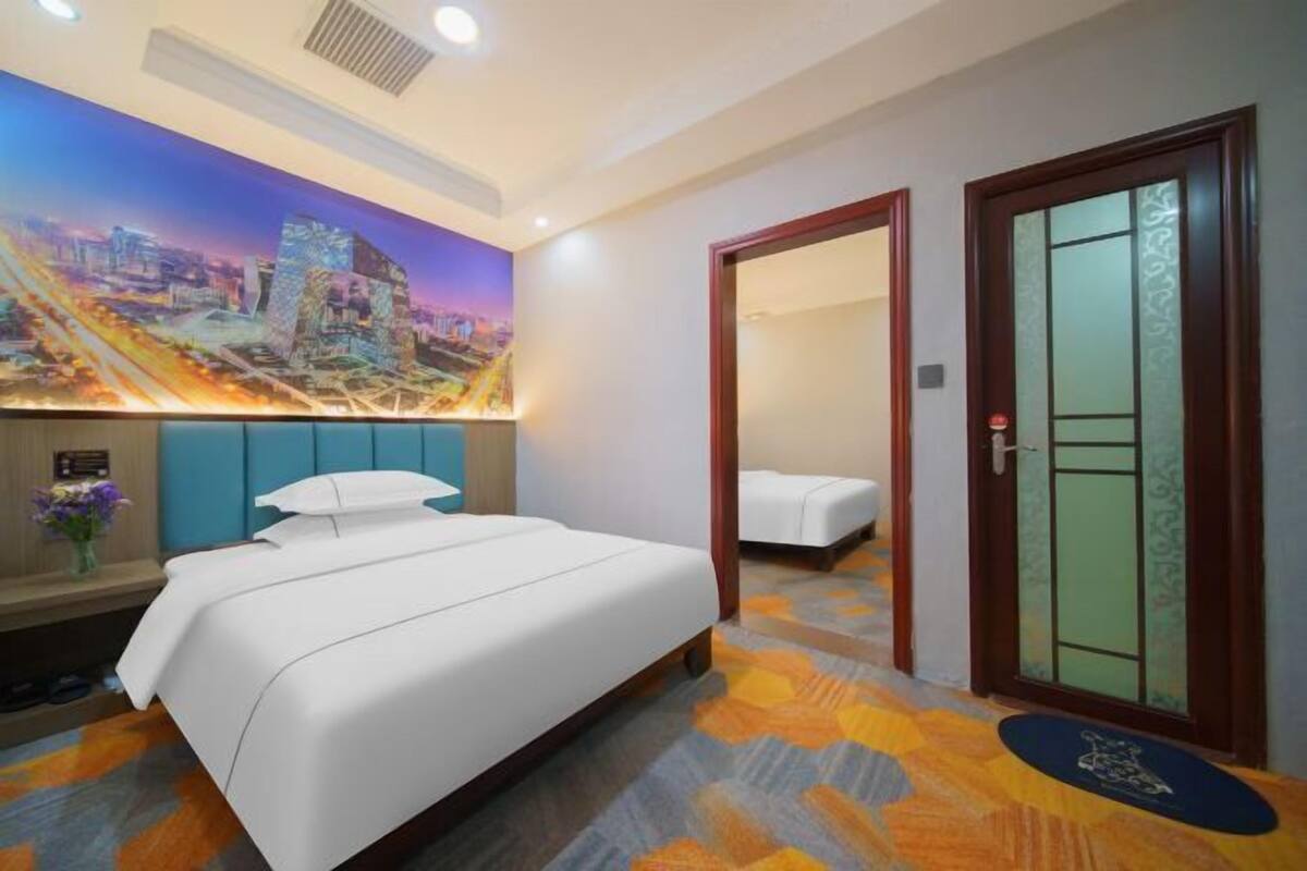 A hotel room with a bed, a nightstand, a wall mural, a door with a glass panel, and a patterned carpet.