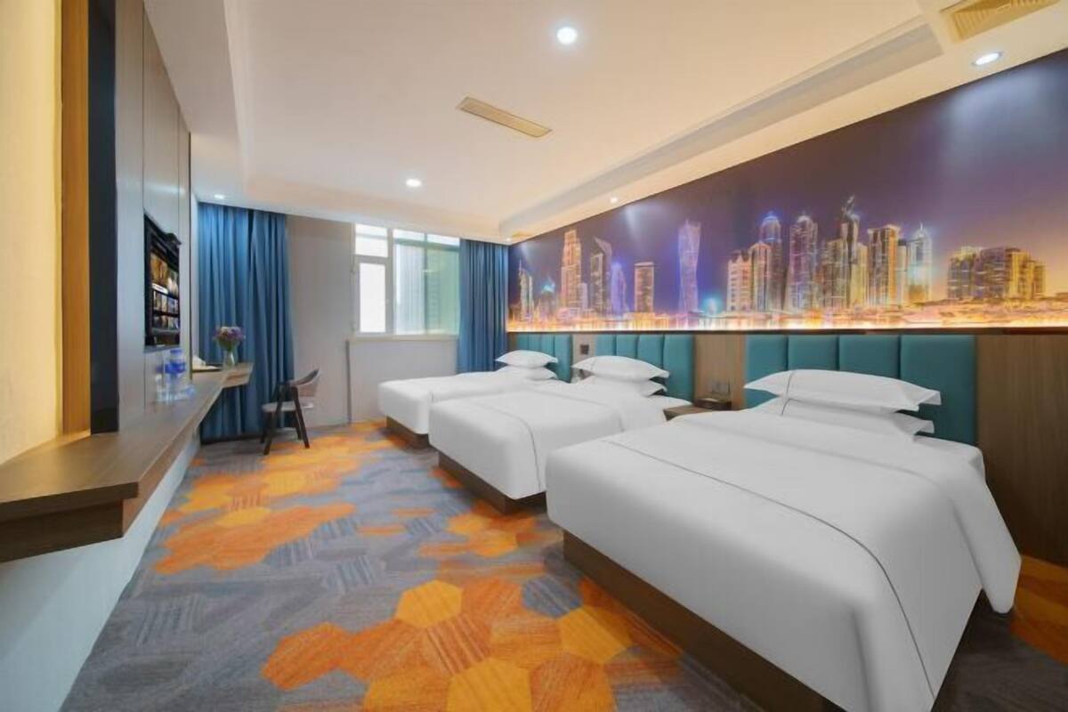 A hotel room with three beds, a cityscape mural, a desk with a chair, and a patterned carpet.