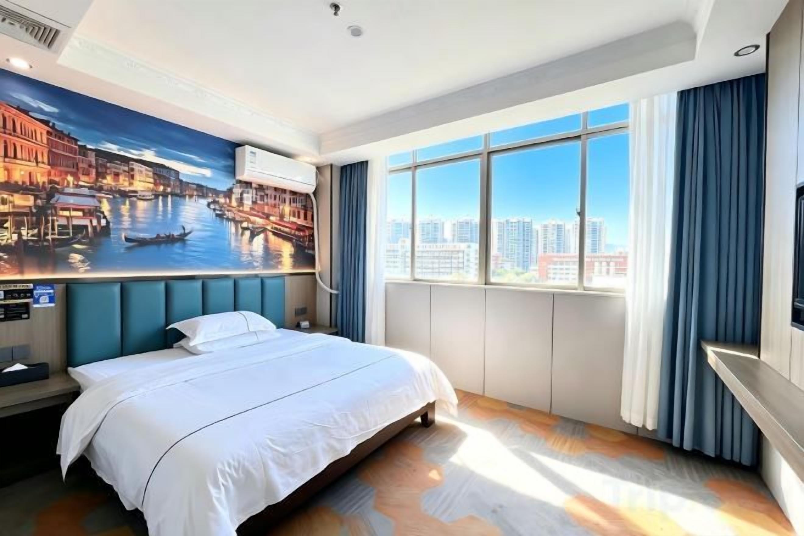 Habitació d'hotel amb un llit, un finestral gran amb cortines, vista a la ciutat i un aire condicionat fixat a la paret.