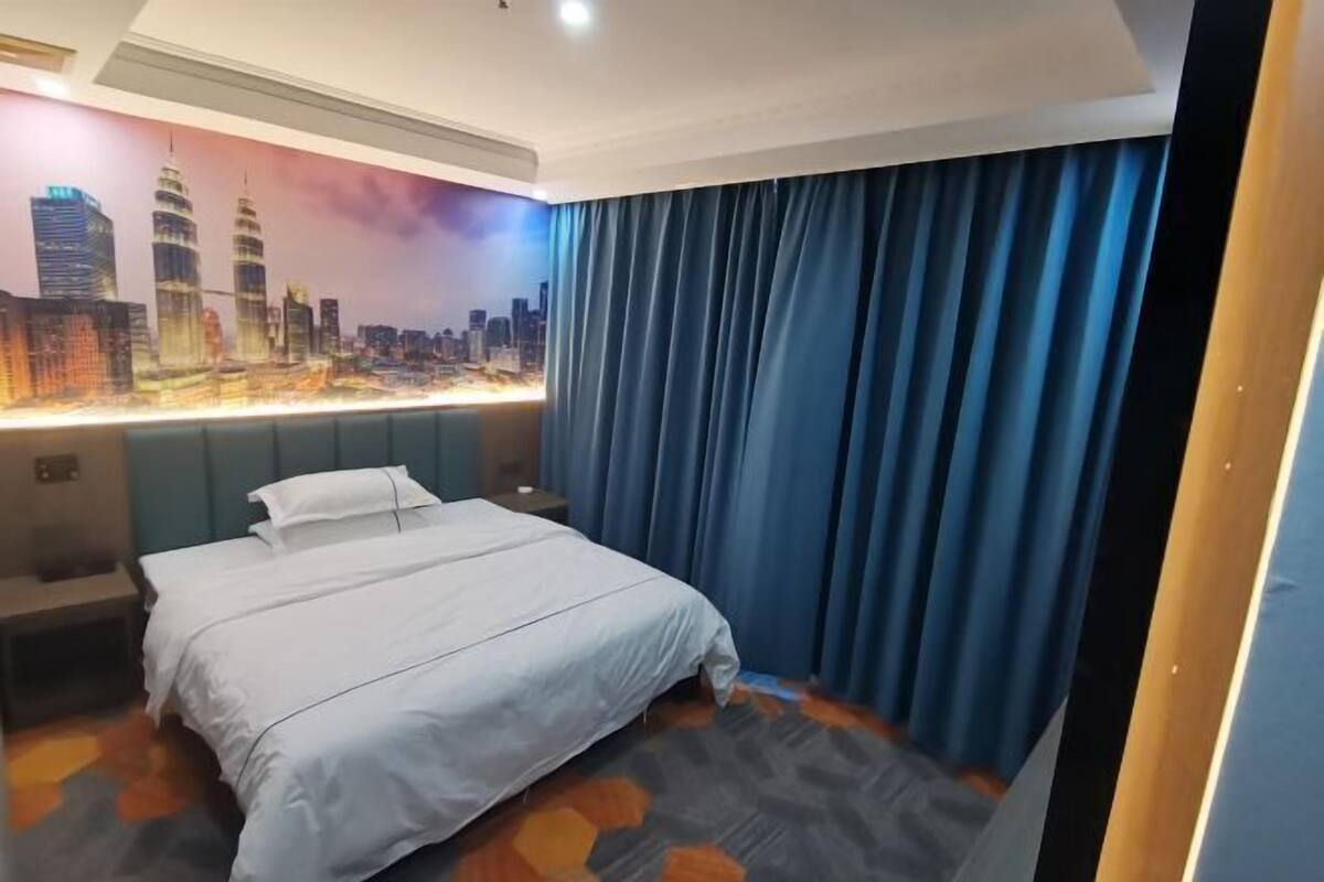 A hotel room with a bed, a nightstand, a wall-mounted light, and a cityscape mural.
