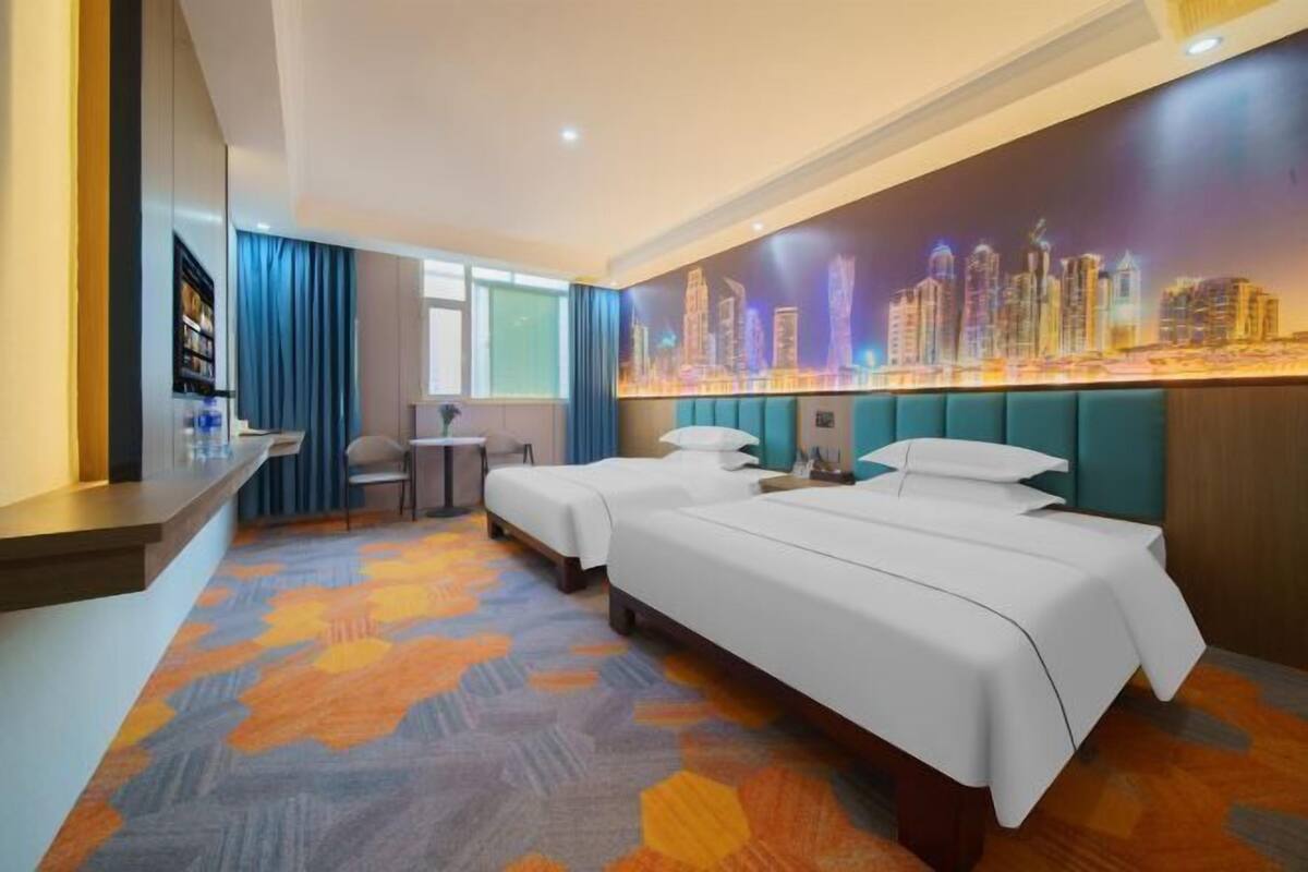 A hotel room with two beds, a desk, a chair, a cityscape mural, and a patterned carpet.