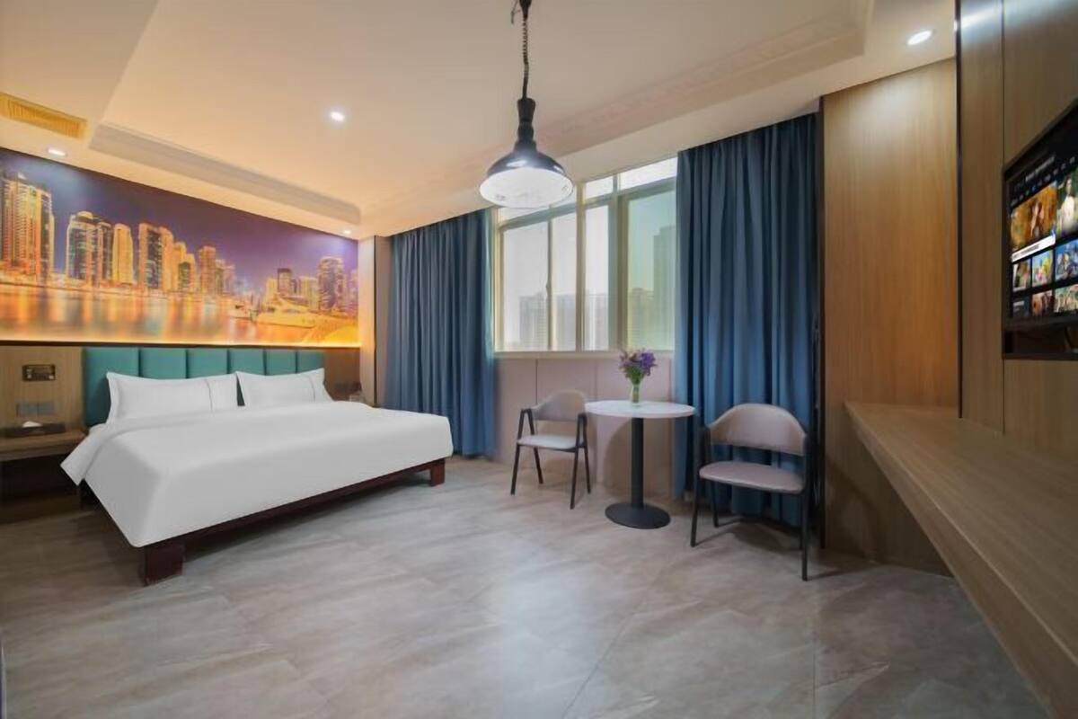 A hotel room with a bed, a small table with chairs, a cityscape mural, and a large window with curtains.