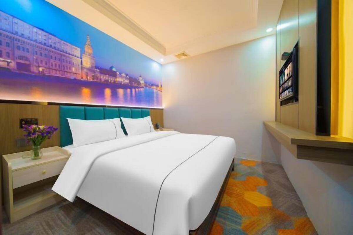 A hotel room with a large bed, a nightstand, a vase of flowers, and a cityscape mural on the wall.