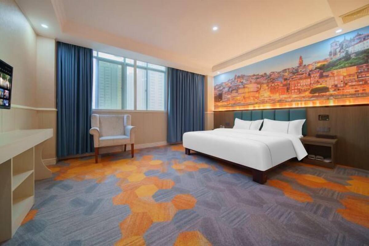 A hotel room with a bed, a chair, a desk, and a large cityscape painting on the wall.