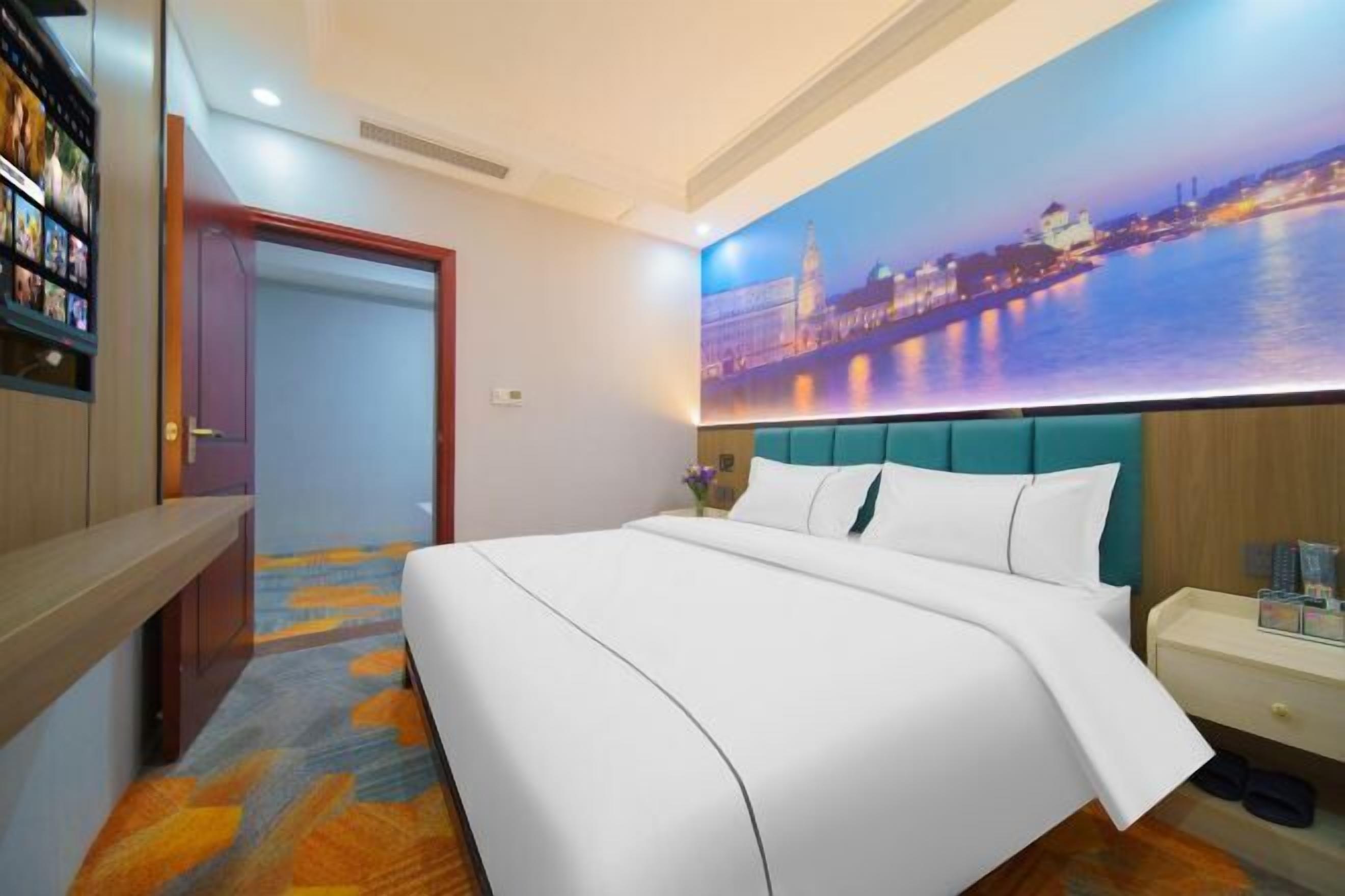 A hotel room with a large bed, a nightstand, a bookshelf, and a cityscape mural on the wall.