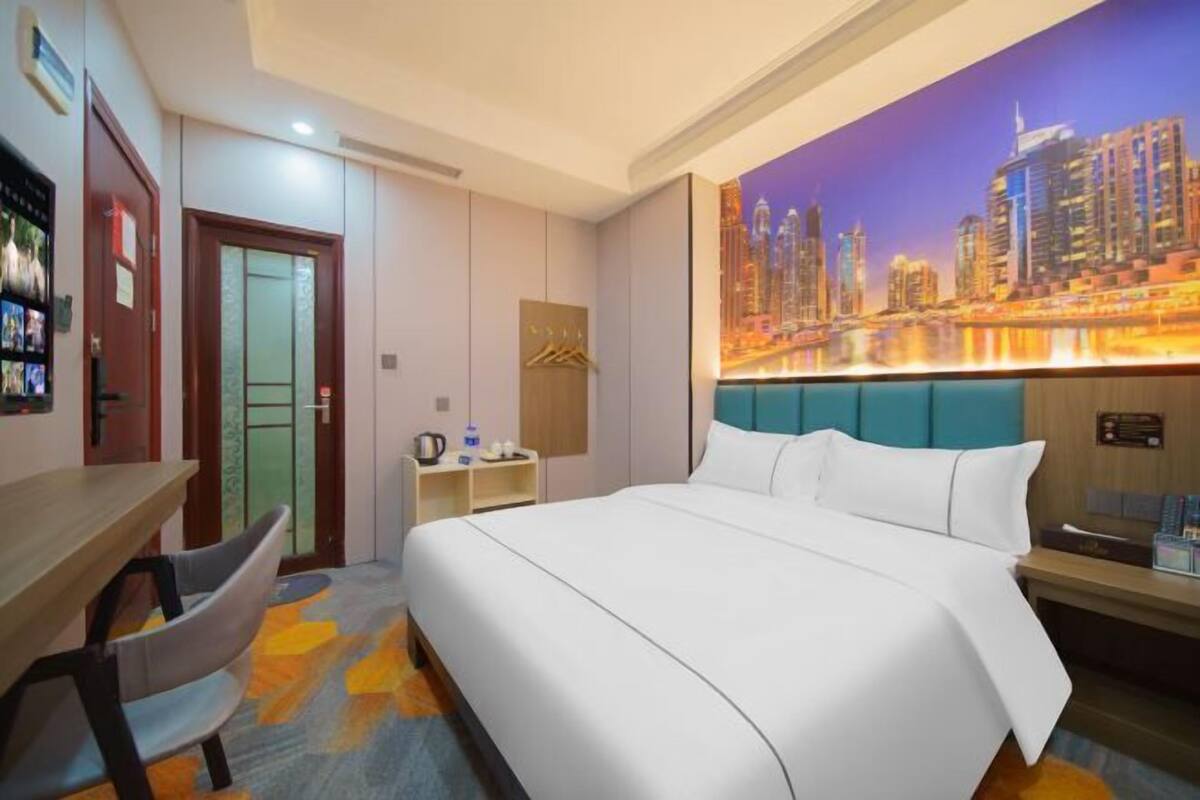 A hotel room with a large bed, a desk with a chair, a small table with a lamp, and a cityscape mural on the wall.