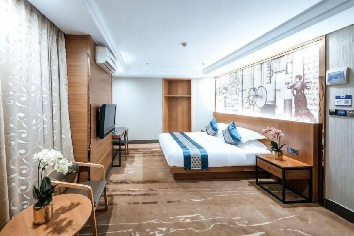 A hotel room with a bed, a desk, a chair, a TV, and a wall with artwork.