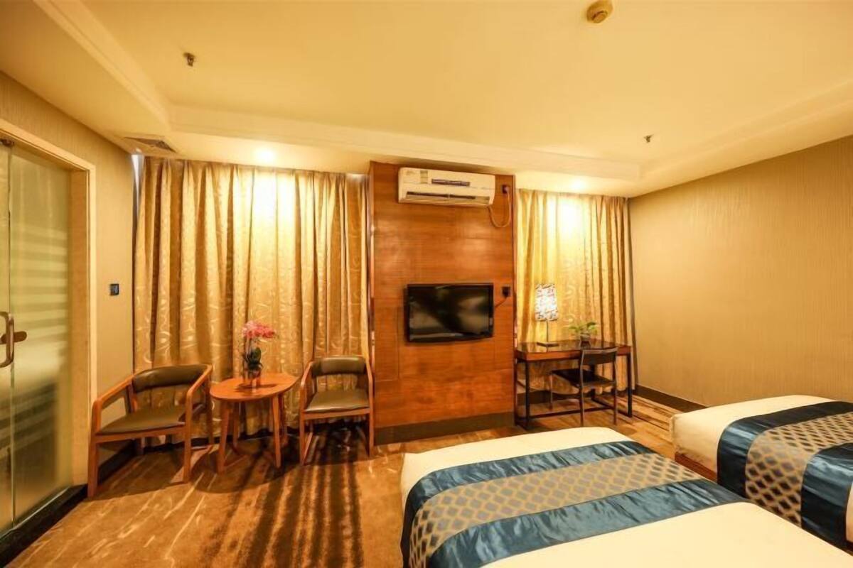 A hotel room with two beds, a TV, an air conditioning unit, a desk with a chair, and a small table with a vase.