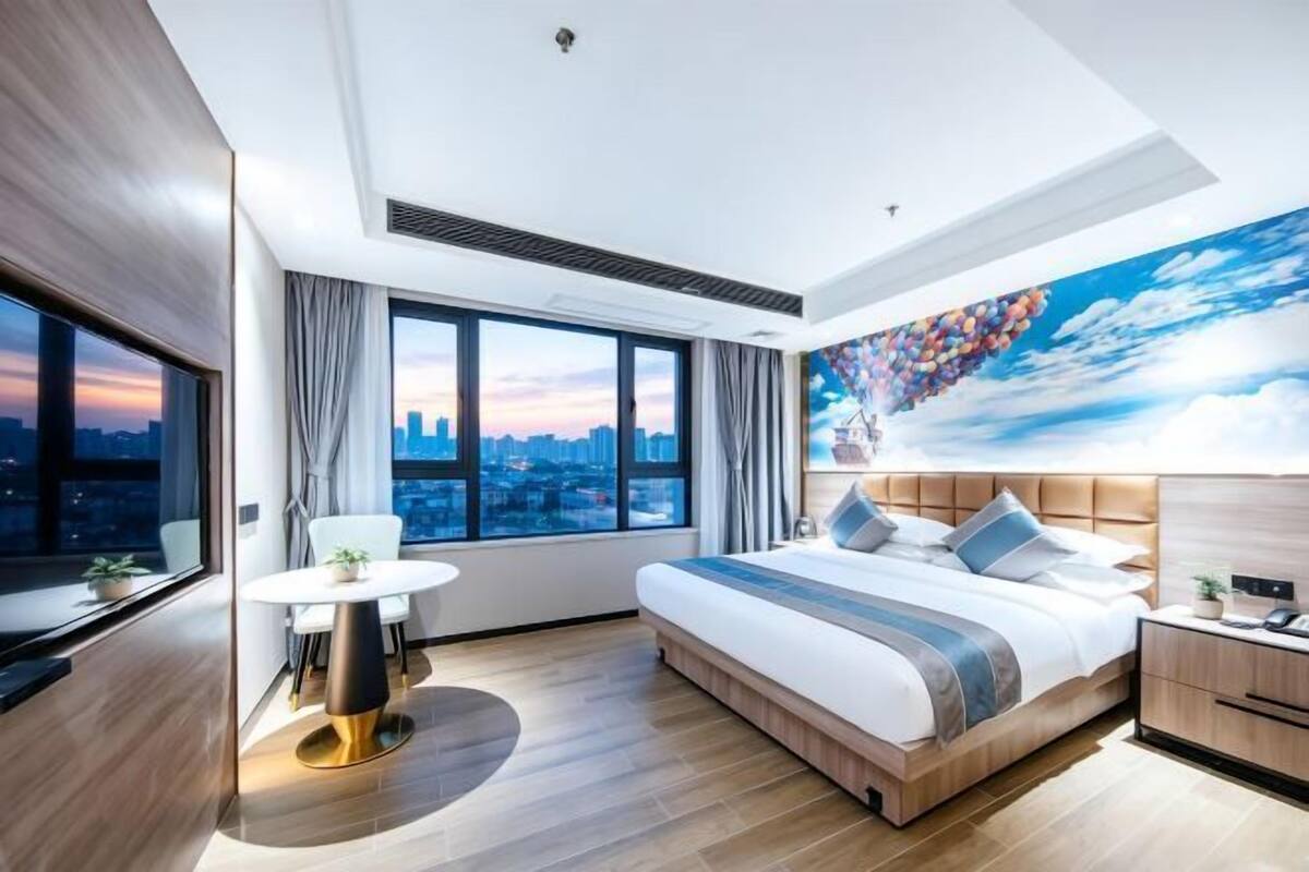 A modern hotel room with a large bed, a round dining table, a city view, and a mural on the wall.