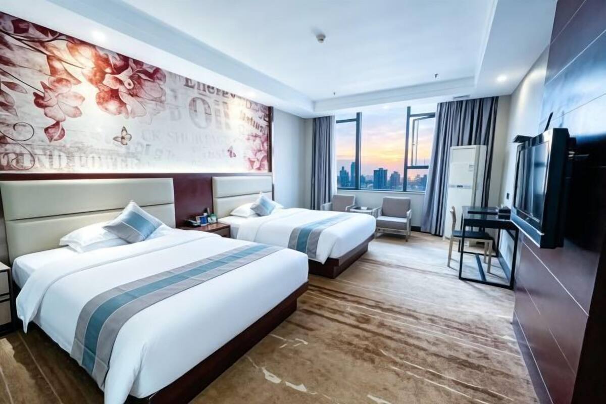 A hotel room with two beds, a desk, a chair, a TV, and a large window with a city view.