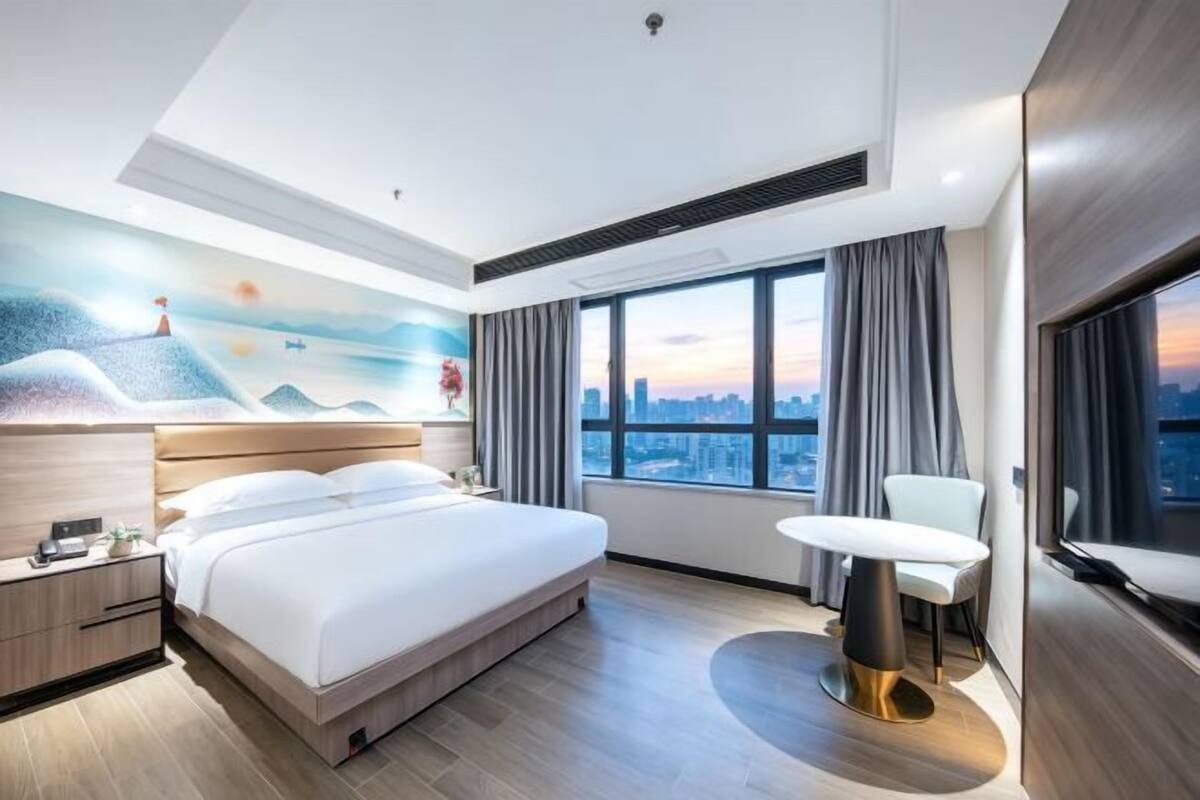 A modern hotel room with a large bed, a city view, a dining table, and a TV.