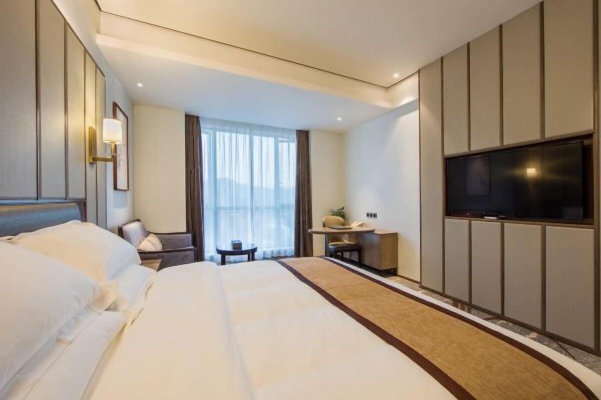 A hotel room with a large bed, a TV mounted on the wall, a desk with a chair, and a window with curtains.