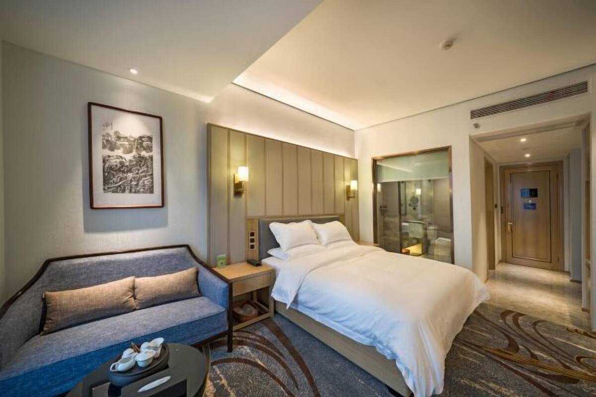 Une chambre d’hôtel comprenant un lit, un canapé, une table de chevet, un tableau encadré, un miroir et un espace douche.