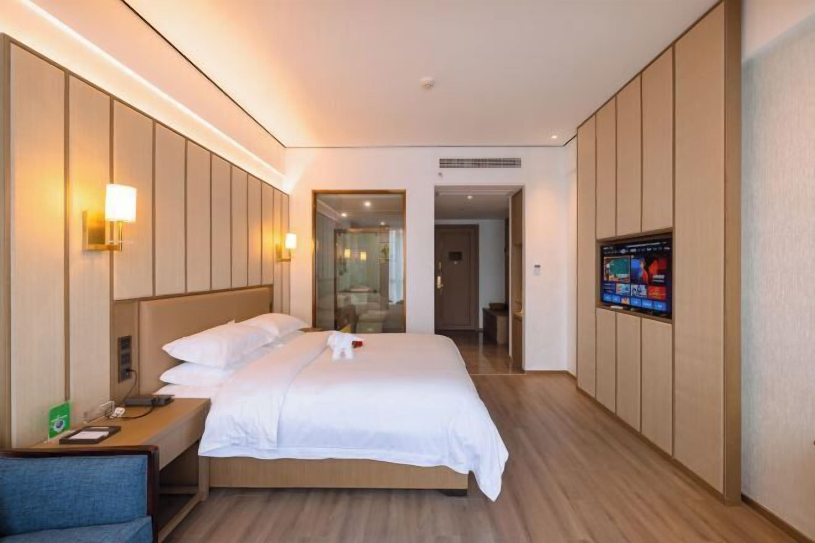 Habitación de hotel con cama, mesita de noche, lámpara, televisor y vista al pasillo.