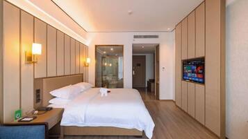 Habitación de hotel con cama, mesita de noche, lámpara, televisor y vista al pasillo.