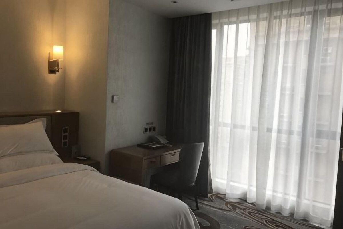 A hotel room with a bed, desk, chair, and a large window with curtains.