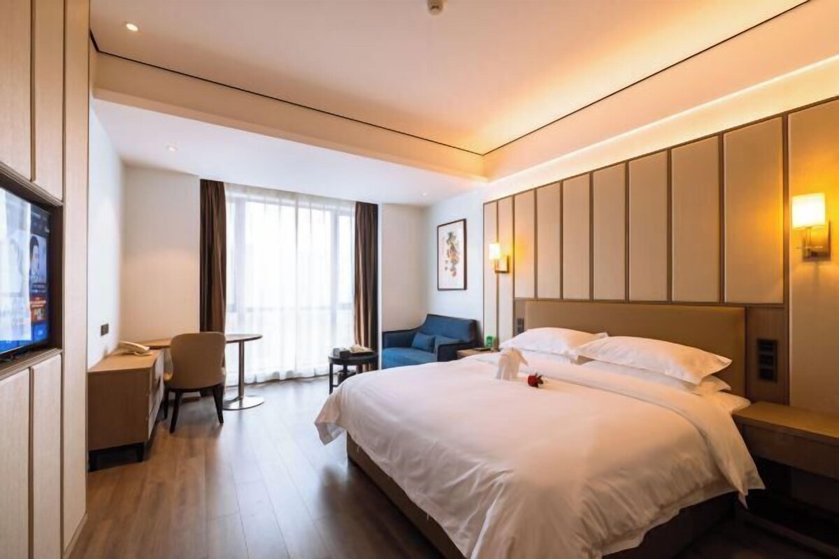 Une chambre d’hôtel avec un grand lit, un bureau, une chaise, une télévision et une fenêtre avec des rideaux.