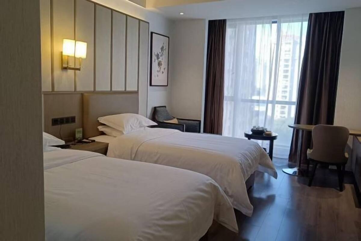 Une chambre d’hôtel avec deux lits, un bureau, une chaise, une petite table, une table de chevet, une lampe fixée au mur et une fenêtre avec des rideaux.