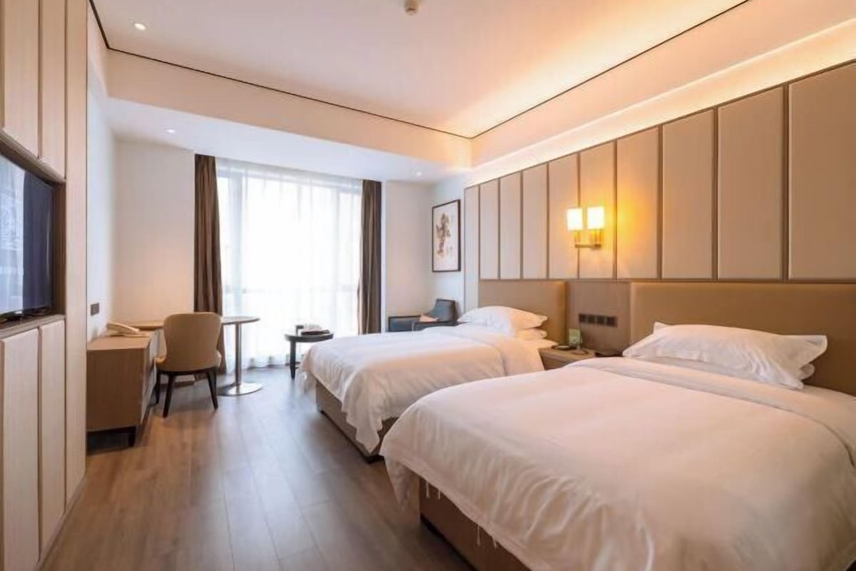 Une chambre d’hôtel avec deux lits, un bureau, une chaise, une petite table et une fenêtre avec des rideaux.