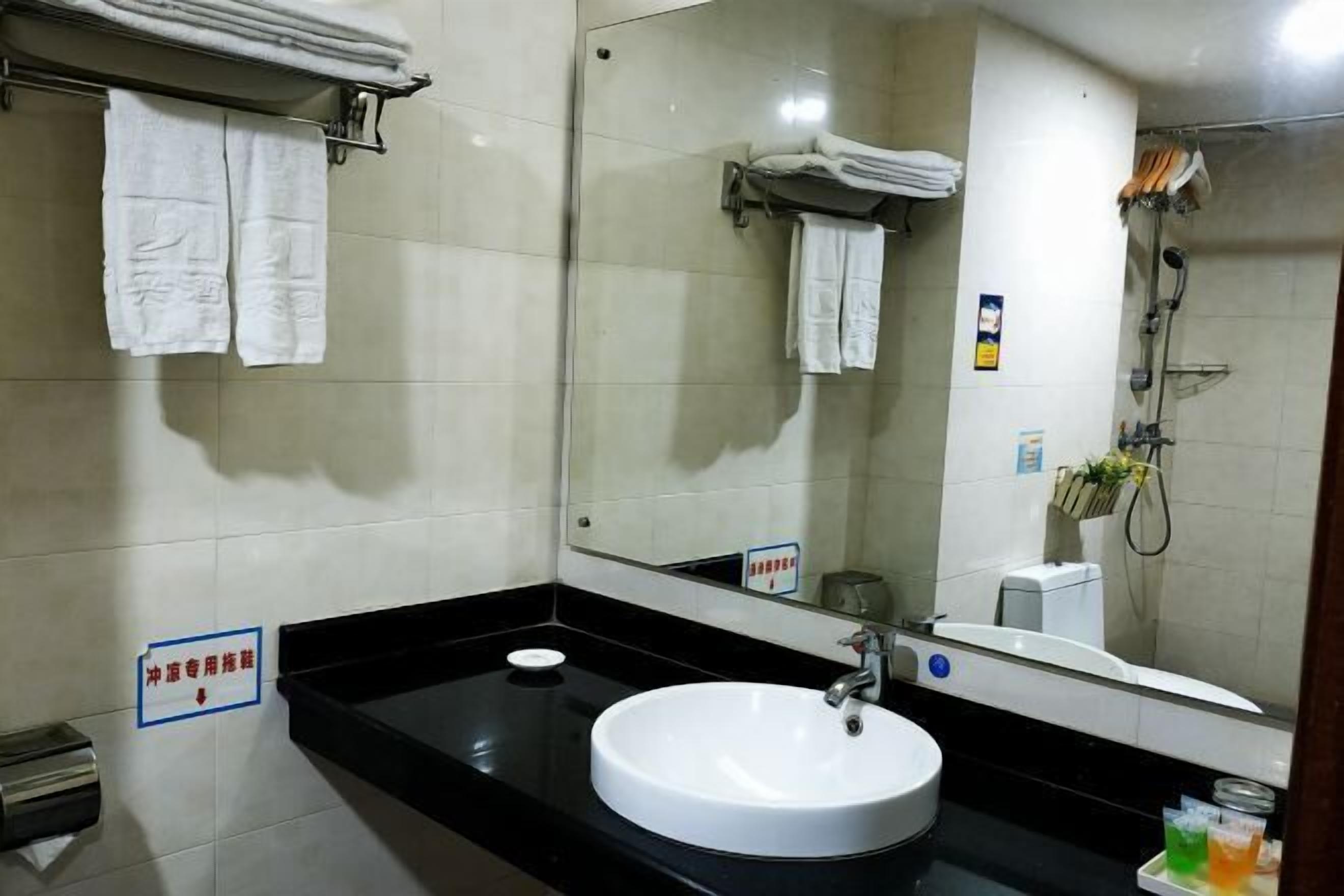 Een badkamer met een wit wastafel, een zwart aanrecht en een spiegel. Handdoeken hangen aan een rek en er is een douche in de achtergrond zichtbaar.