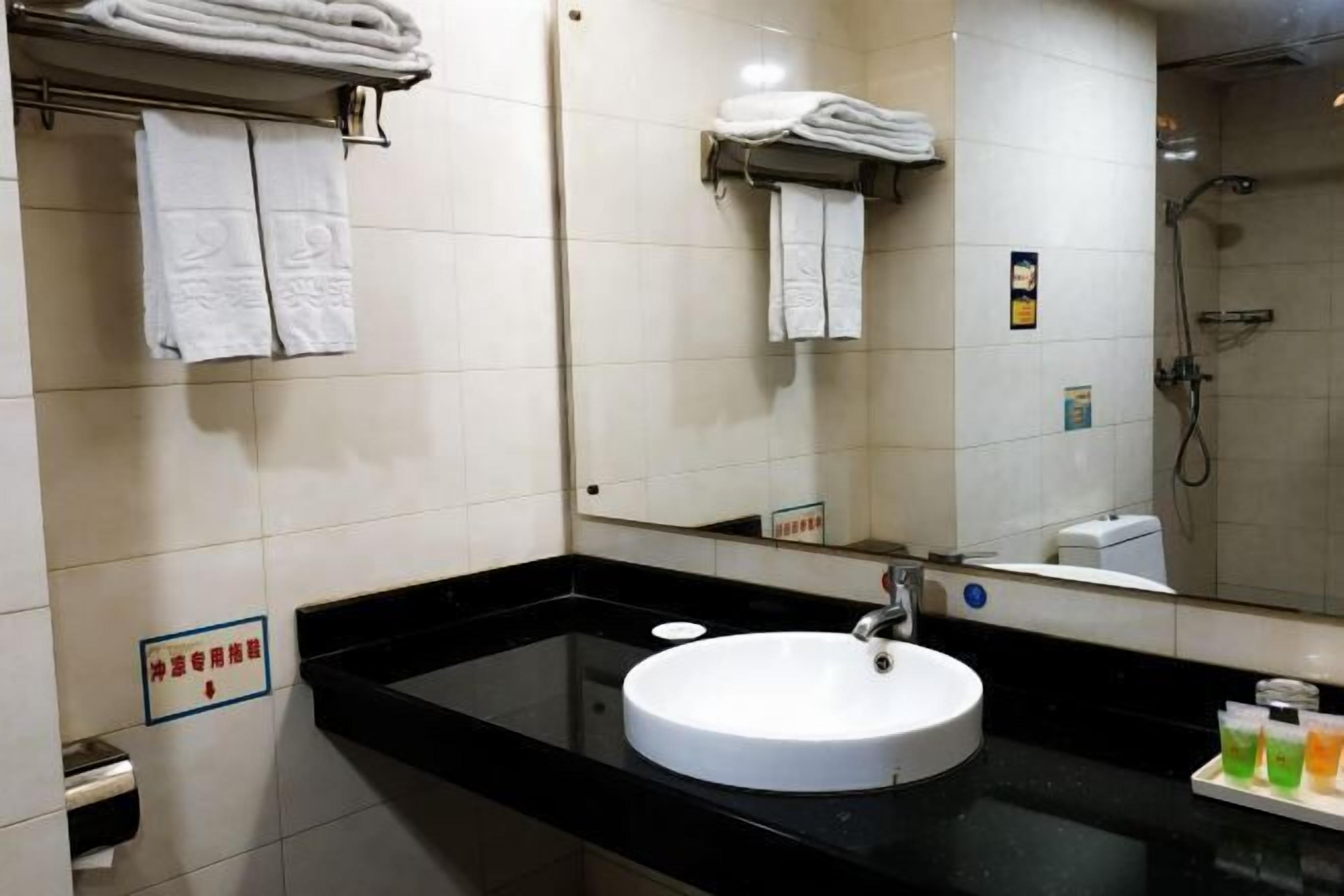 Un baño con un lavamanos blanco, encimera negra y un espejo. Hay toallas en un perchero y una ducha visible al fondo.