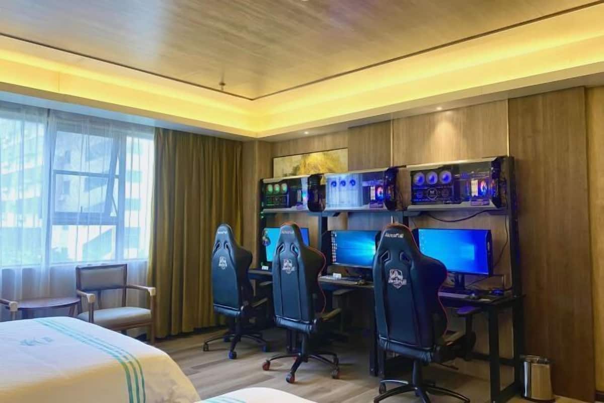 A hotel room with a bed, three gaming chairs, two gaming stations, and a large window with curtains.
