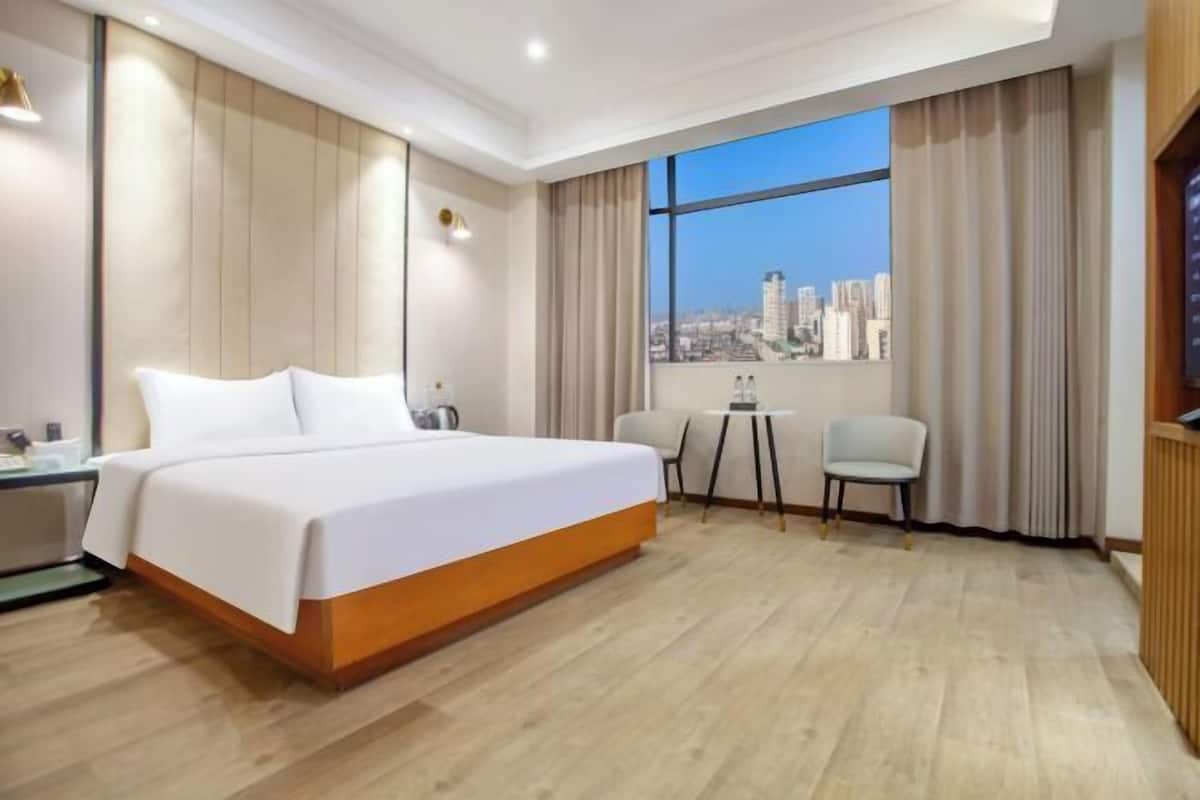 A hotel room with a large bed, a small table, a chair, and a view of the city through a large window.