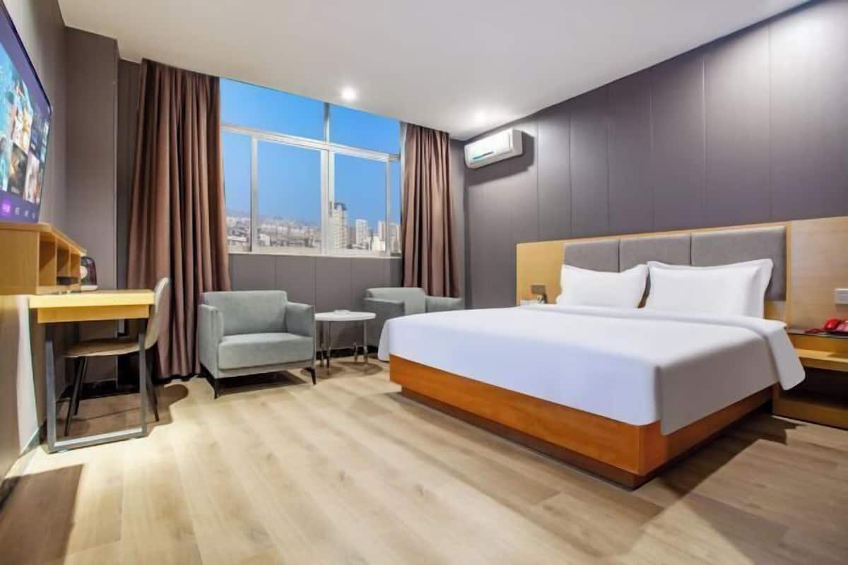 A hotel room with a large bed, a desk, a chair, a small table, and a window with a city view.