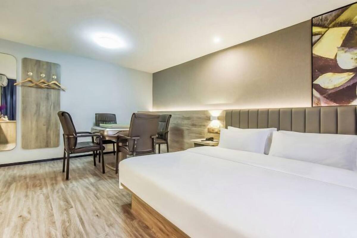A hotel room with a bed, two chairs, a desk, a lamp, and a wall-mounted artwork.