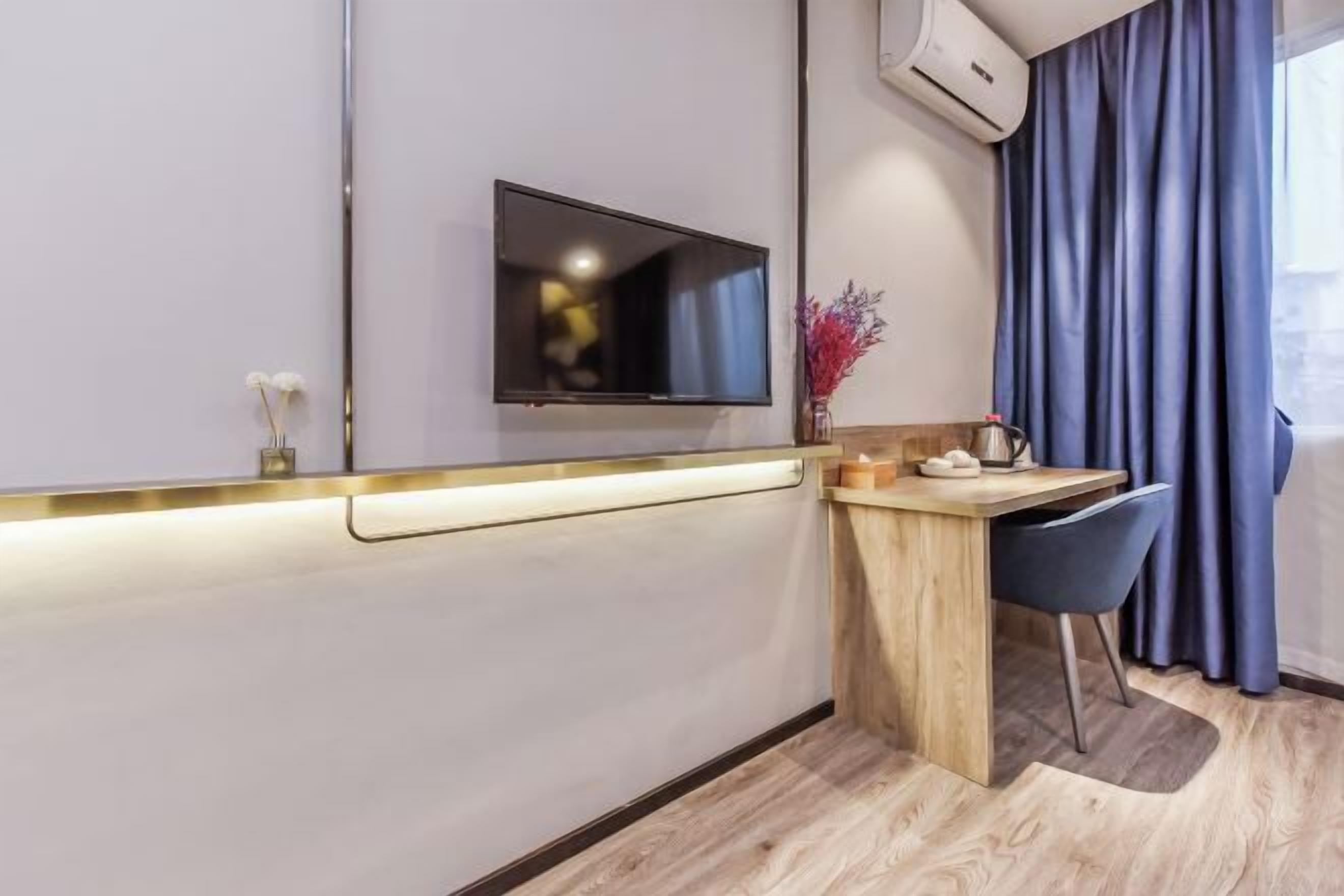 Quarto de hotel moderno com televisão de tela plana na parede, escrivaninha de madeira com cadeira, vaso pequeno com flores e janela com cortinas azuis.