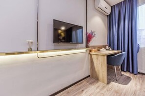 Habitació d'hotel moderna amb televisió de pantalla plana penjada a la paret, escriptori de fusta amb una cadira, un vas petit amb flors i una finestra amb cortines blaves.
