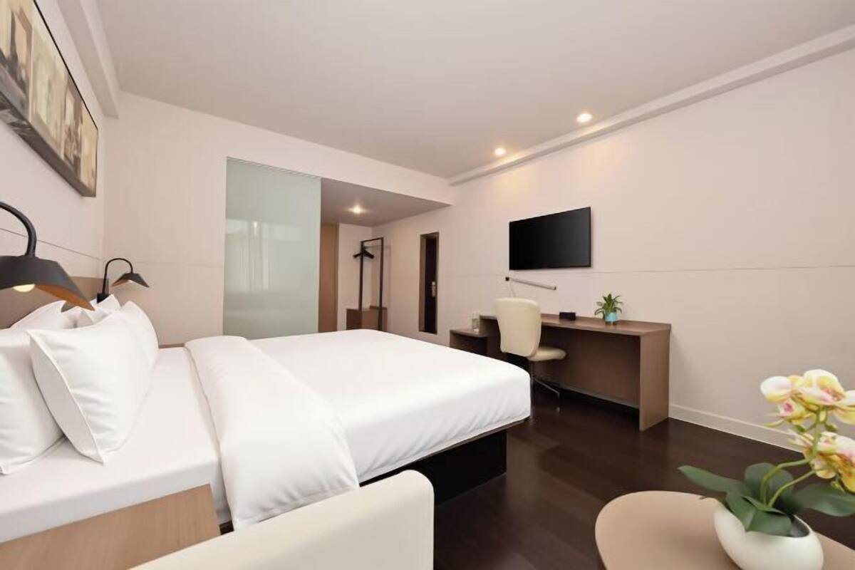 A hotel room with a bed, desk, chair, TV, and a plant.