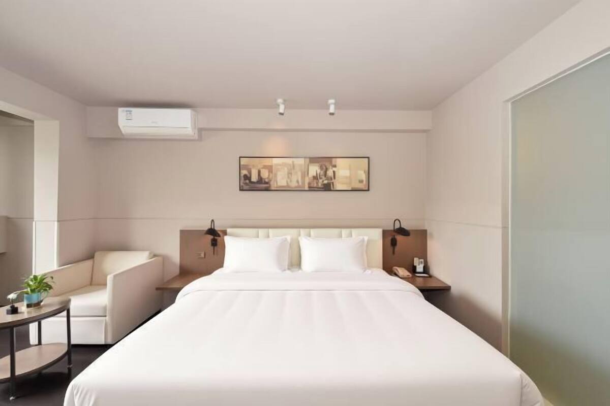 A hotel room with a bed, a chair, a small table with a plant, a wall-mounted air conditioner, and a framed picture above the bed.