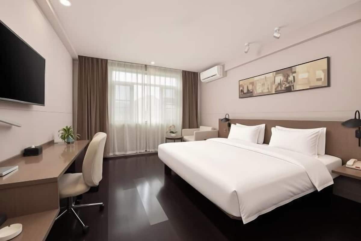 A hotel room with a bed, desk, chair, TV, artwork, and an air conditioning unit.