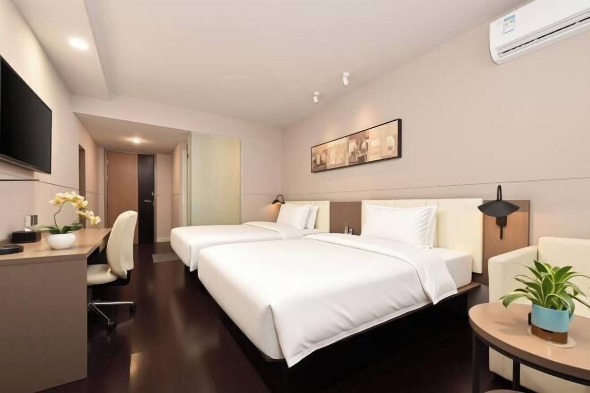 A hotel room with two beds, a desk, a chair, a TV, and a plant.