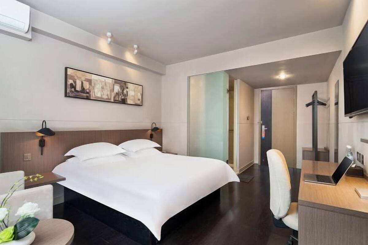 A hotel room with a bed, a desk with a laptop, a chair, a wall-mounted TV, and a framed picture.