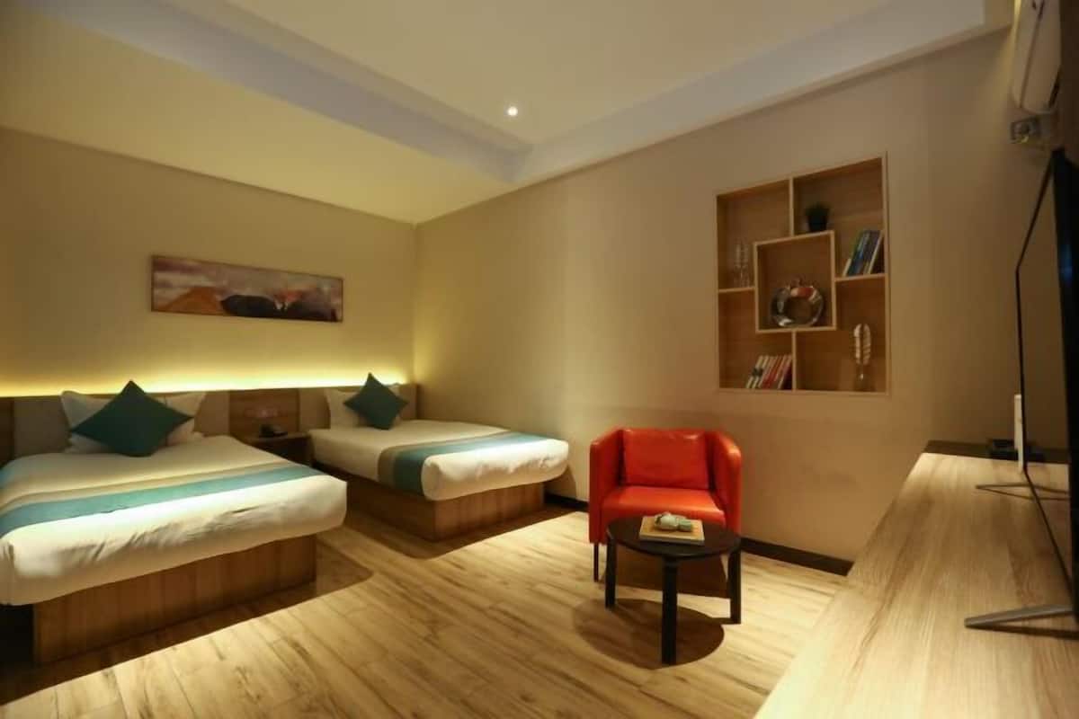Quarto de hotel com duas camas, uma poltrona vermelha, uma mesinha e uma estante de livros e objetos decorativos.
